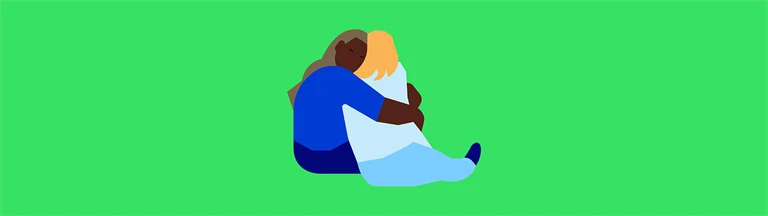 Kvinna tröstar en annan med en kram, de sitter på golvet.